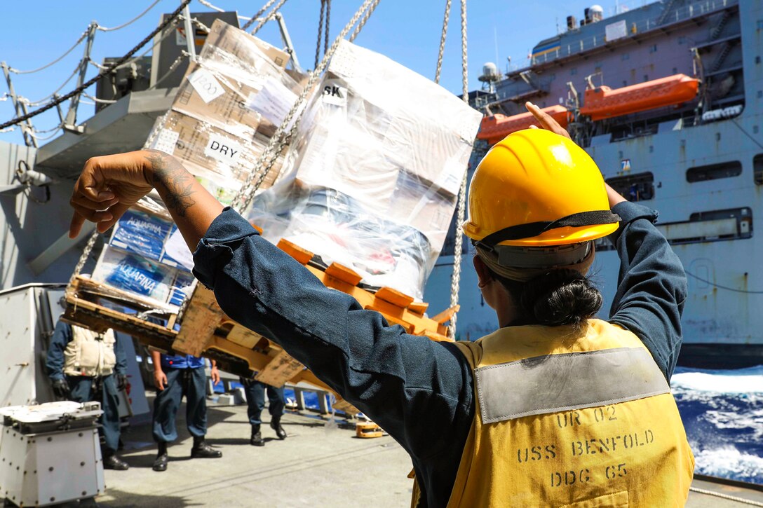 A sailor signals to an operator to lower a pallet onto a ship.