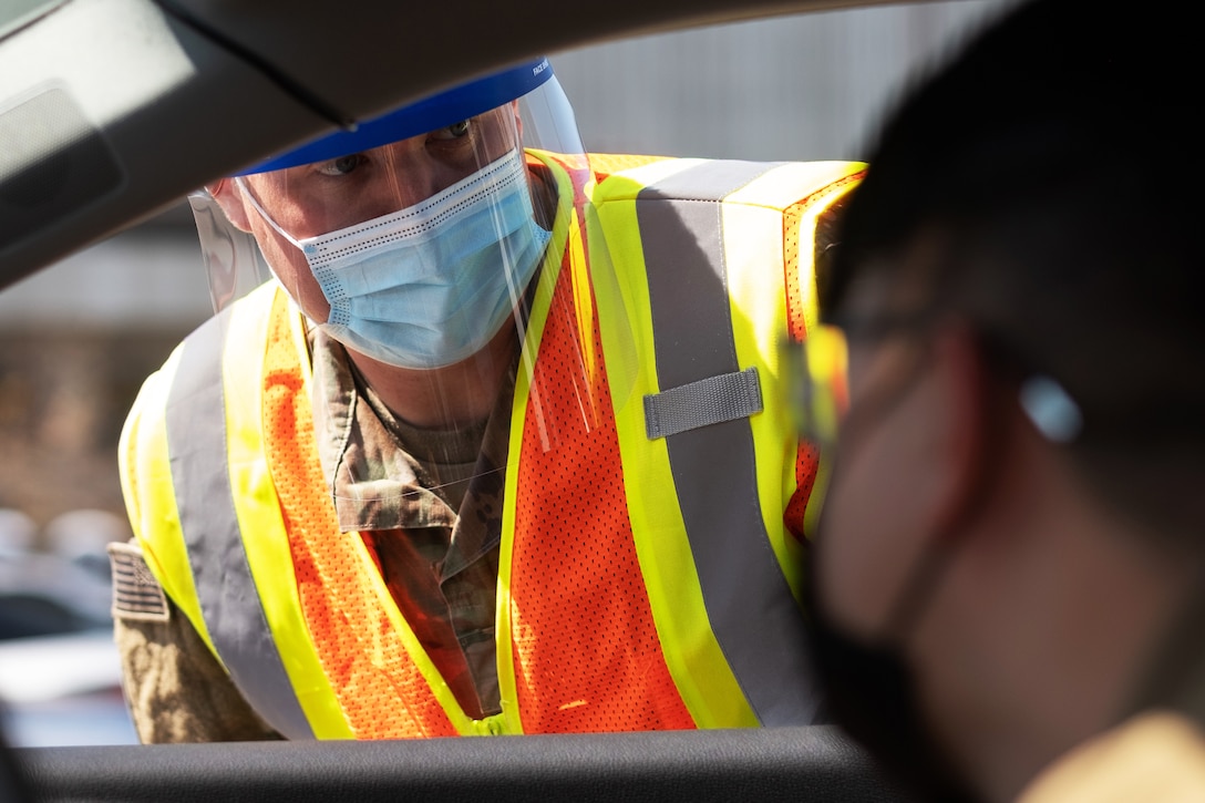 A soldier wearing a face mask leans over into a car window to answer a question.