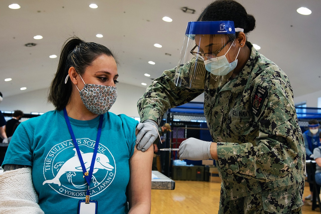 A sailor wearing a face mask and gloves gives a woman seated in a chair wearing a face mask a vaccine.