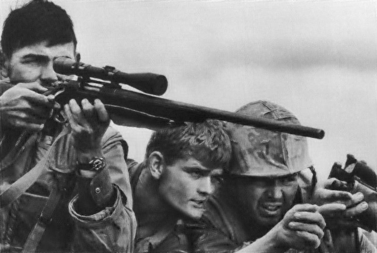 A sniper aims a rifle while two soldiers discuss a target.
