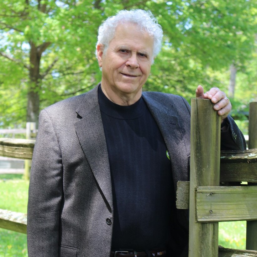 A white-haired man stands by a gatepost and looks at the camera.