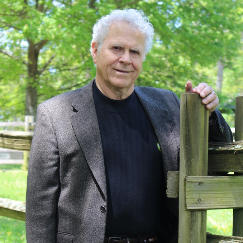 A white-haired man stands by a gatepost and looks at the camera.