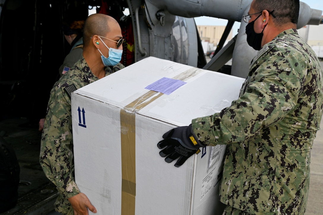 Two seamen both wearing face masks and one wearing gloves load a box of vaccines onto a helicopter.