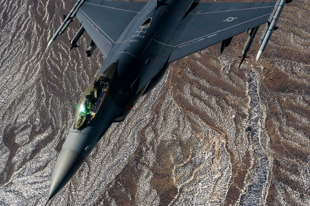 An aircraft flies over dry, brownish terrain.