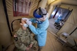 Two uniformed service members are in a tent. One wears PPE and a faceshield, while they use a nasal swab to collect a COVID-19 sample from the other Service member in uniform.