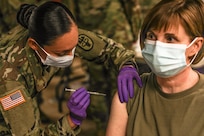 Two masked women service members are in the photo. One servicemember is giving the other a COVID-19 vaccination in her upper arm.