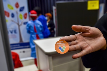 In the foreground a hand holding an orange sticker that says "I got my vaccine". In the background there are three medical personnel wearing personal protective equipment.