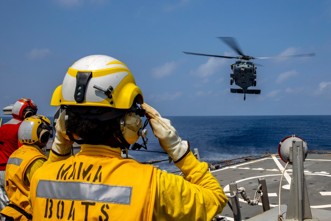 A sailor signal an airborne helicopter from the deck of a ship.