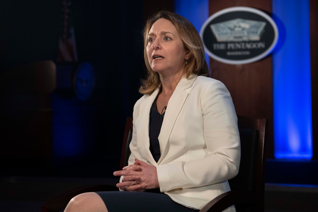 A seated woman in business dress speaks to a man across from her. The Pentagon logo is on a wall behind her.