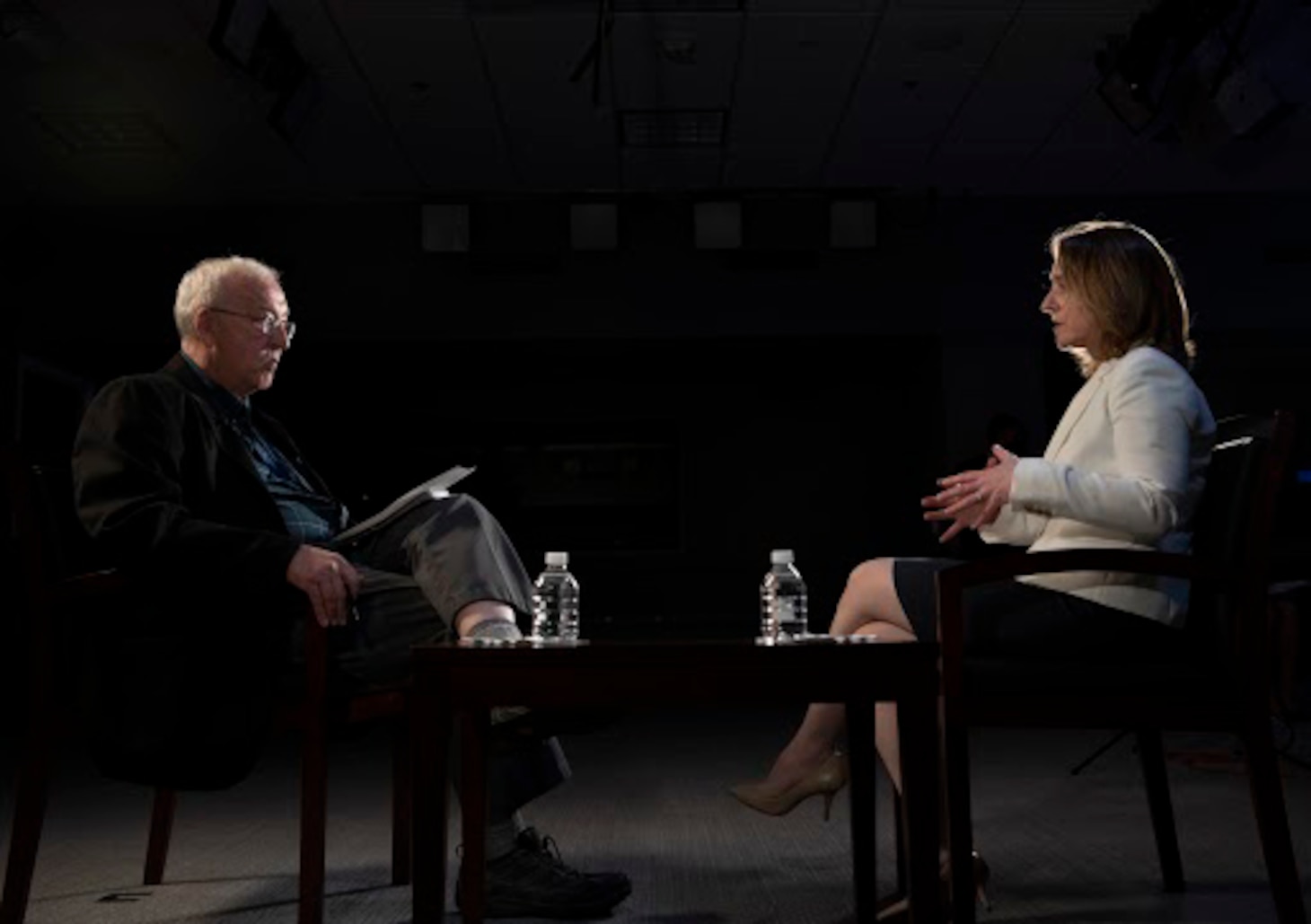 A man and a woman in business attire talk while seated in a darkened room.