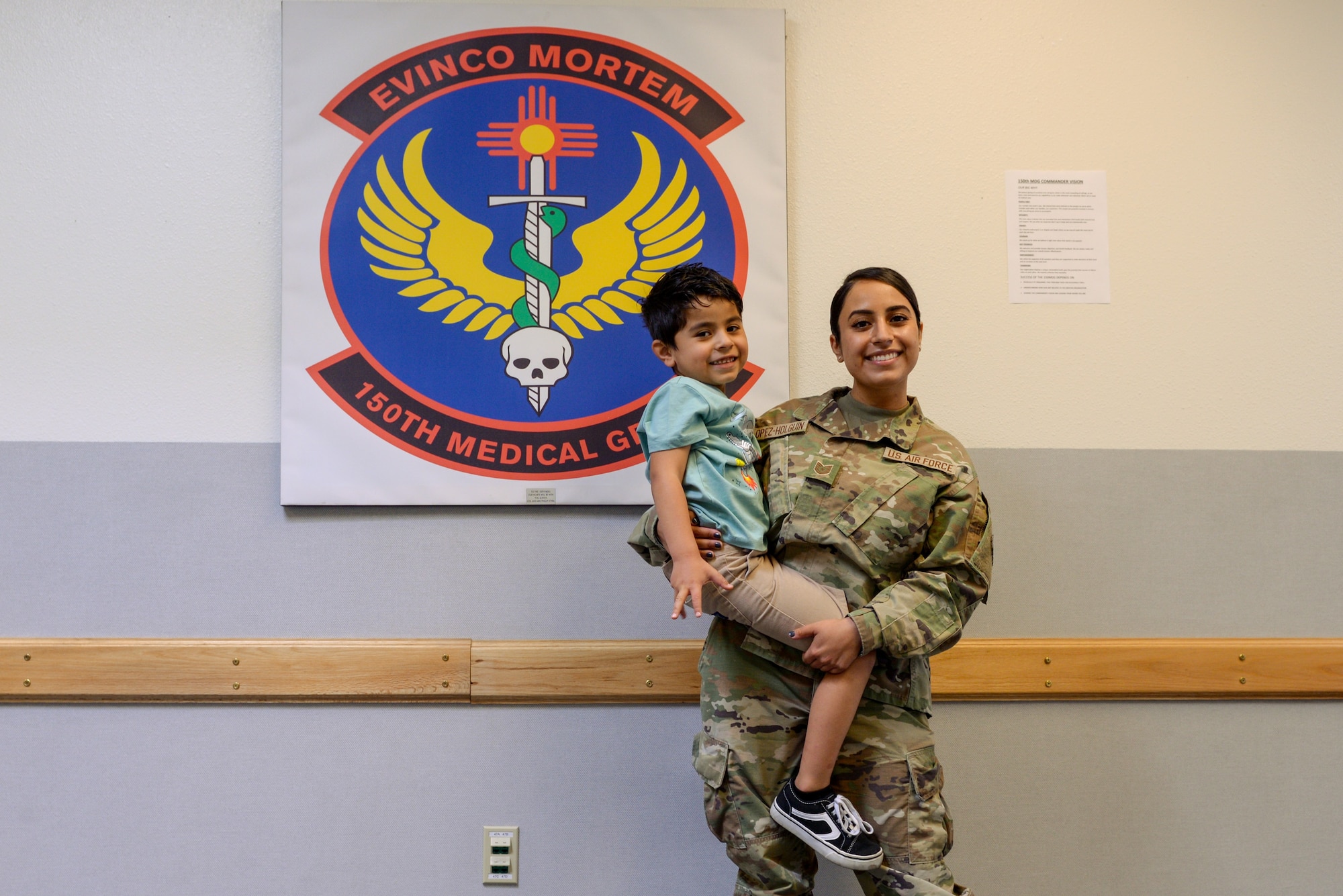 Tech. Sgt. Raven Lopez stands in front of the 150th medical group logo while holding her son in the lobby of the 150th medical building