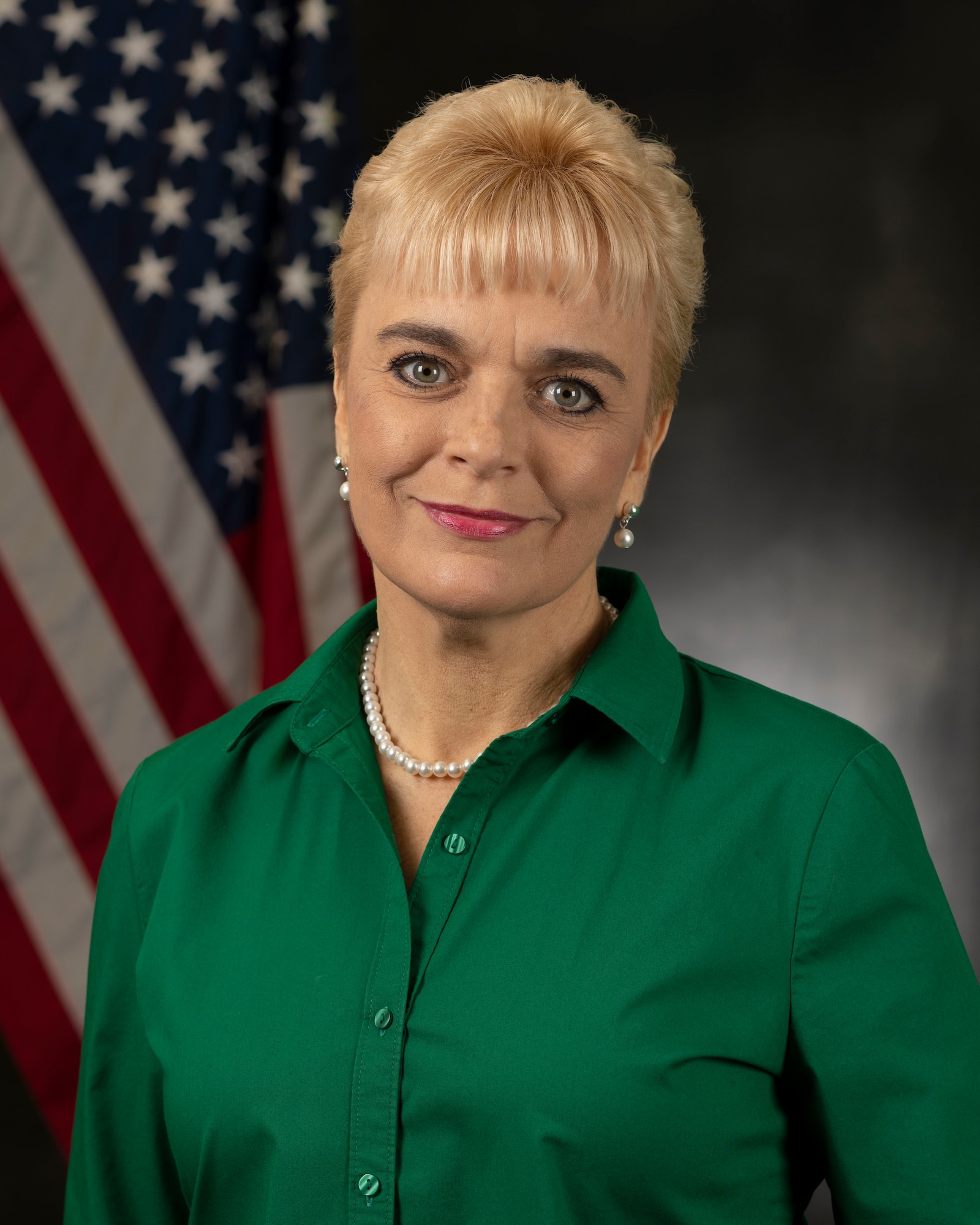 A blonde woman in a green shirt smiles and poses in front of an American Flag and grey backdrop.