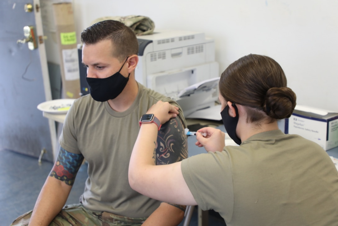 A seated man in a protective mask receives a shot from a crouching woman also wearing protective equipment.