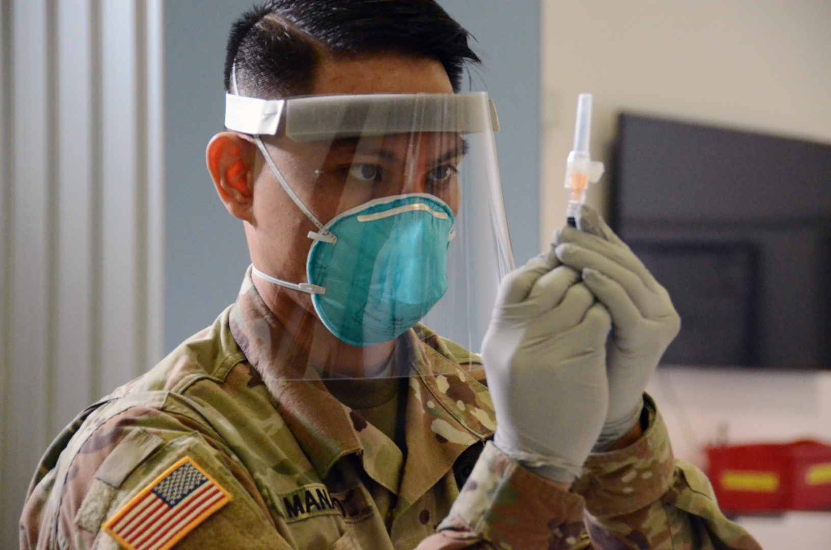 A man in a military operational camo pattern uniform holds a syringe while wearing personal protective equipment.