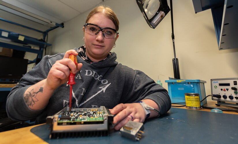 Photo of woman, surrounded by tools, she sits at a work bench to repair a power supply unit.