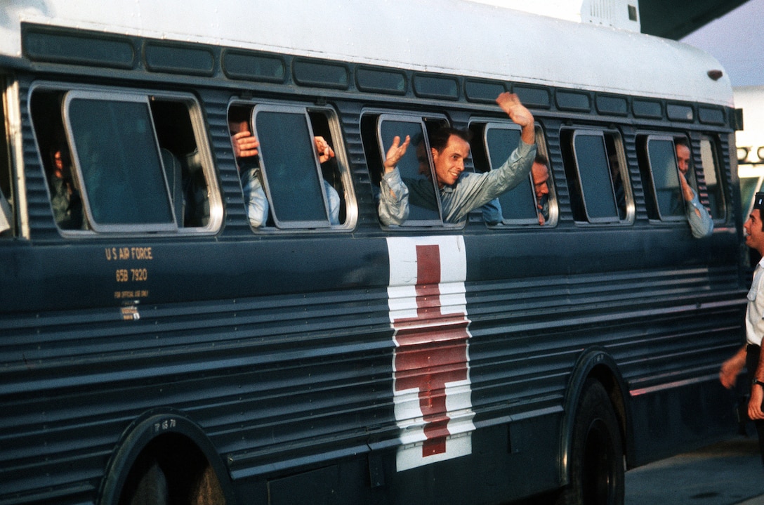 People wave and peek out the windows of a bus with a Red Cross painted on it.