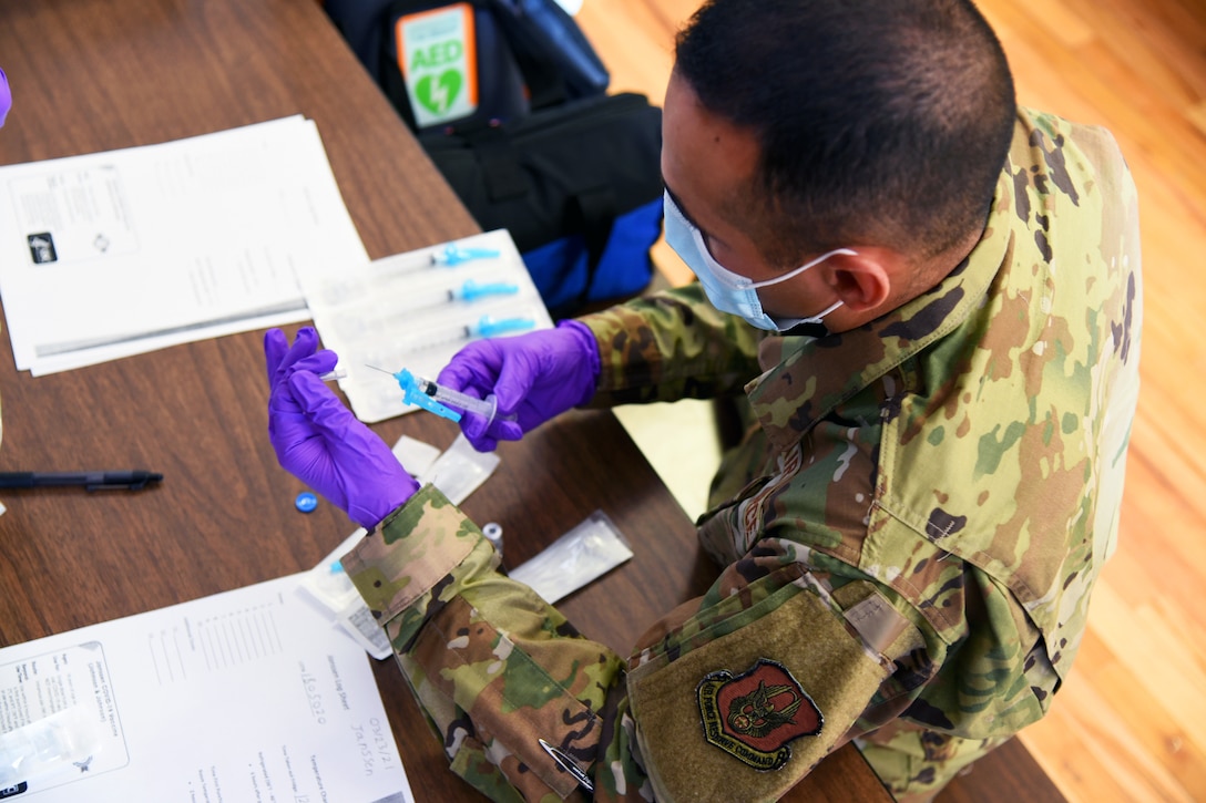 A soldier wearing a face mask and gloves holds a syringe that is being inserted into a vial and filled with a vaccine.