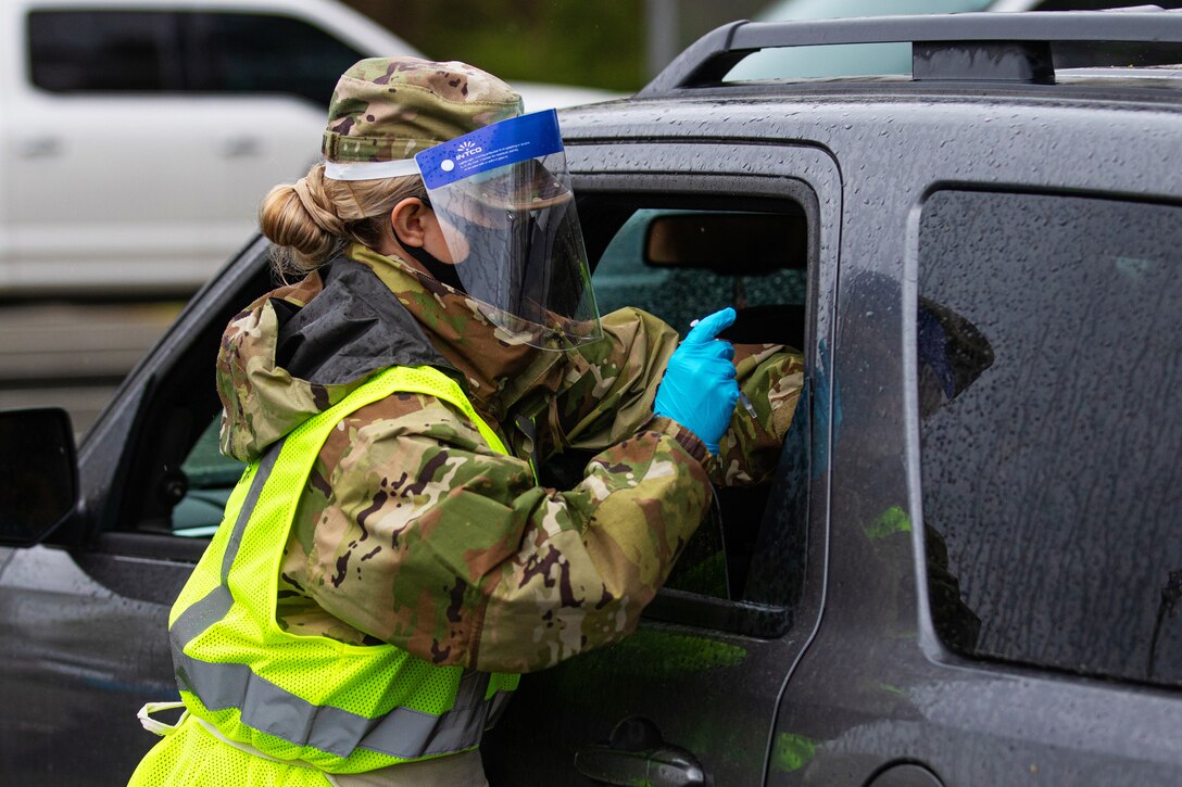 A soldier wearing a face mask and gloves vaccinates a resident whose sitting in their vehicle.
