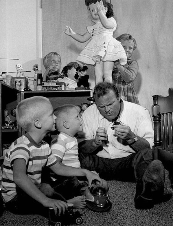 A man sits on the floor playing with two little boys as two little girls play with a large doll and other toys behind the man.