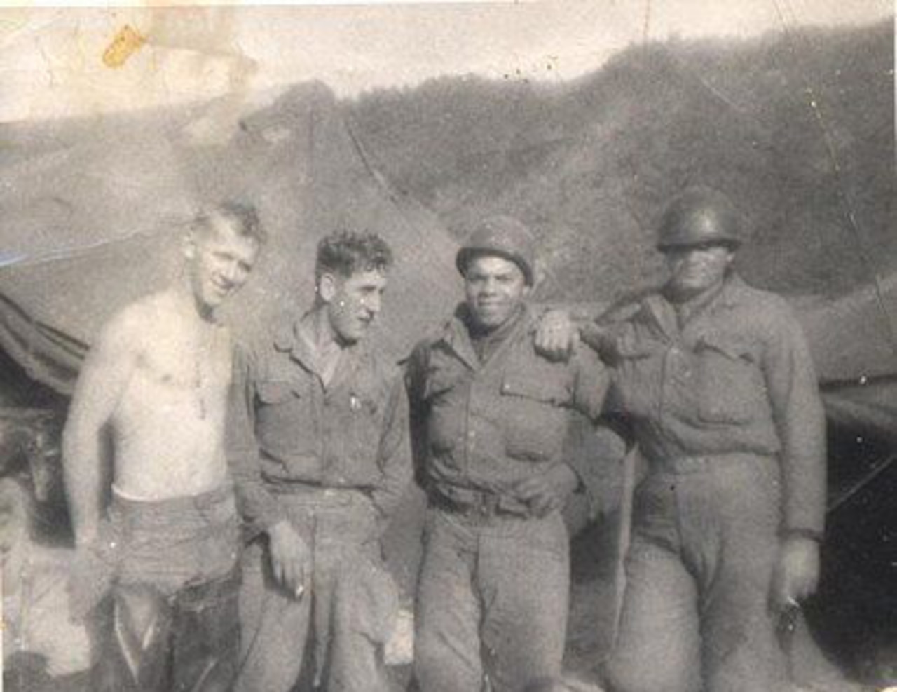 Four soldiers pose for a photo; mountains and tents are in the background.