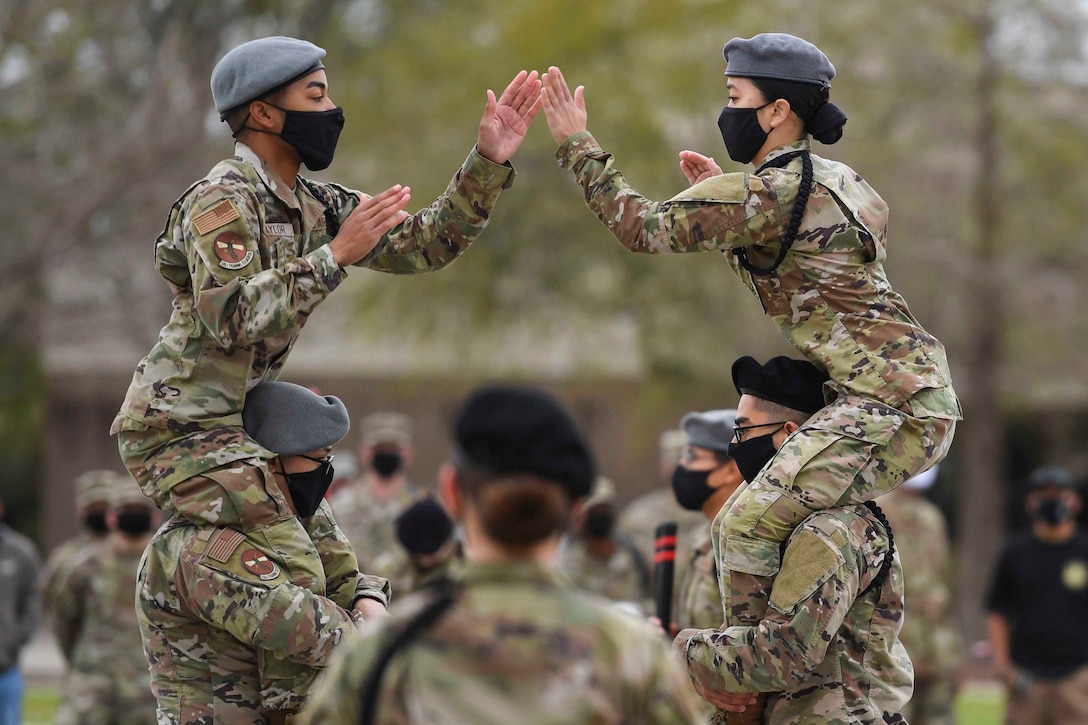 Two airmen sit on the shoulders of two other airmen while clapping hands.