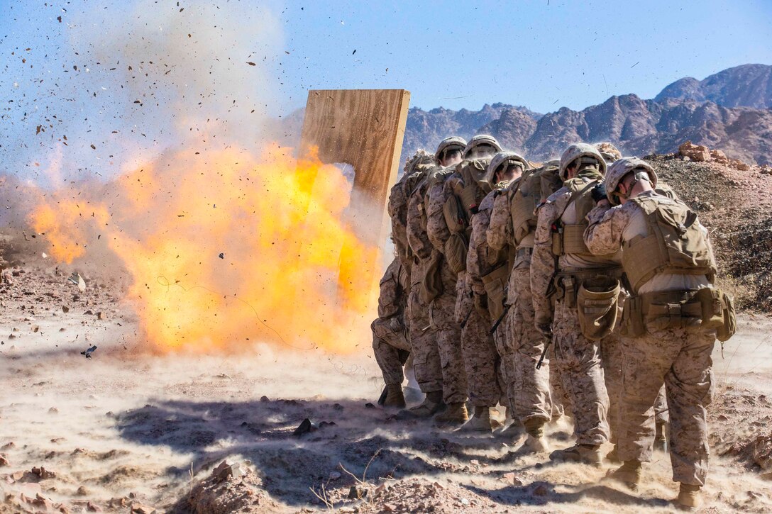 Marines stand huddled together in a line as an explosive device detonates.