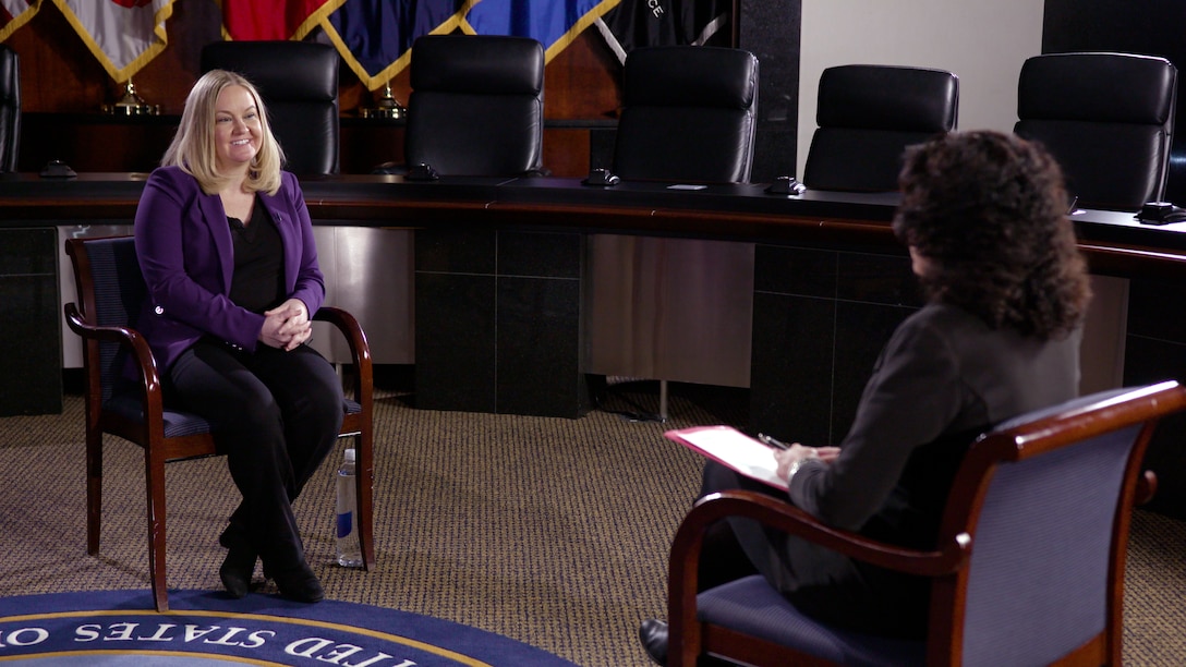 Woman with blond hair in a black shirt and purple sweater sits in a char talking with a woman whose back is facing the camera.