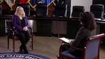 Woman with blond hair in a black shirt and purple sweater sits in a char talking with a woman whose back is facing the camera.