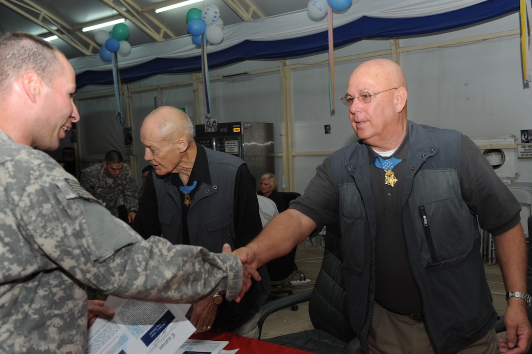 A soldier shakes hands with a man wearing a medal who's standing behind a table.