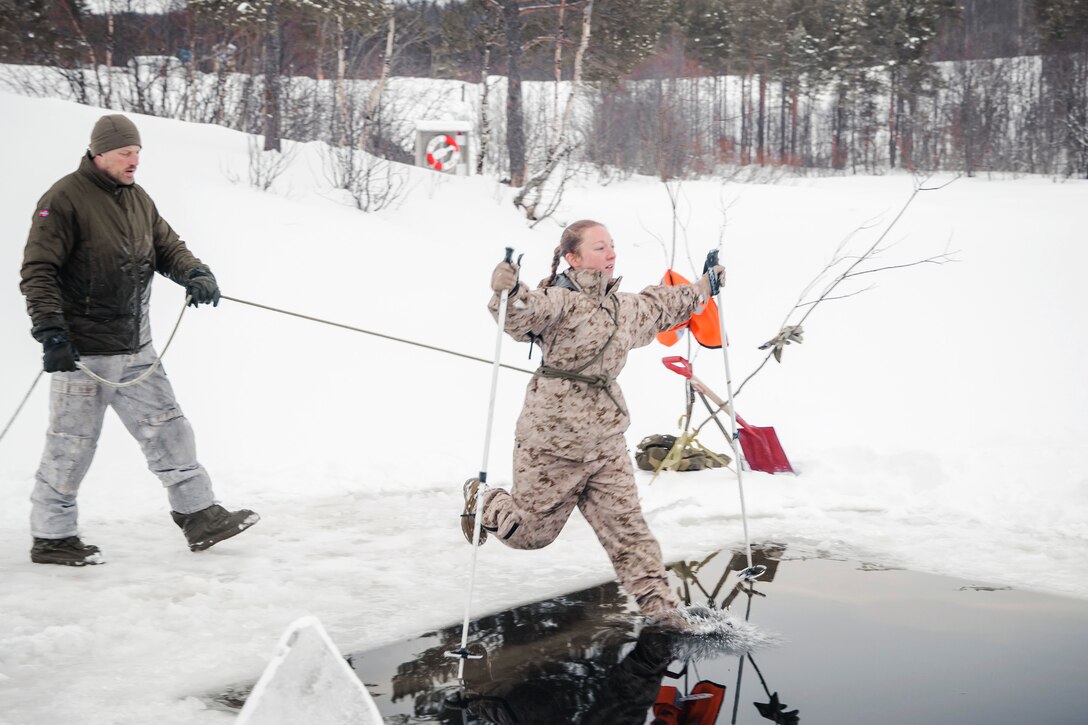 A service member holds a rope connected to a Marine who is jumping into freezing water surrounded by snow.