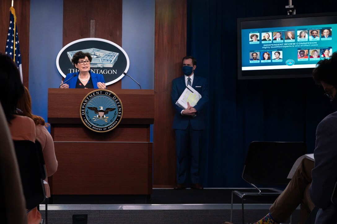 A woman stands at a lectern. A man stands behind her to the left. On a television screen at the rear are the faces of multiple individuals. A sign on the lectern reads "Department of Defense."