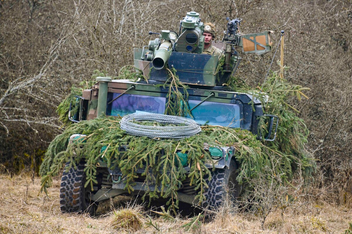 A soldier travels in a military vehicle camouflaged by tree branches.