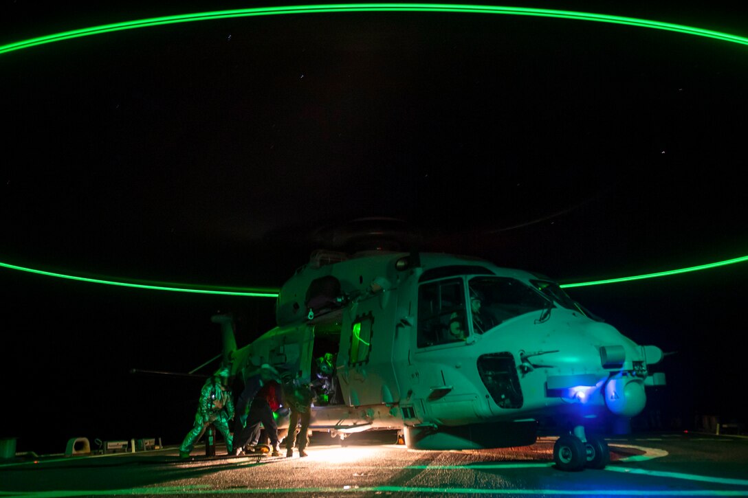 Sailors aboard a ship refuel a helicopter illuminated by neon lights.