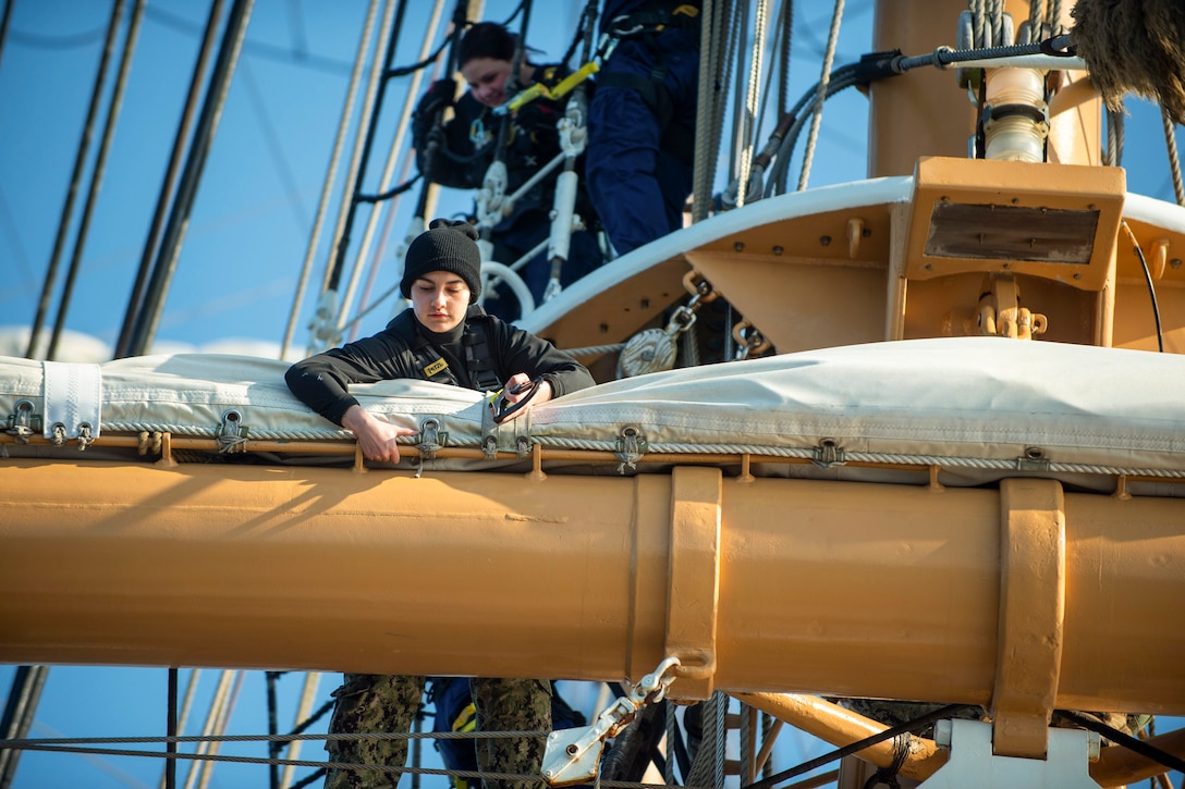 A sailor climbs on the forward course yard of a ship during training.