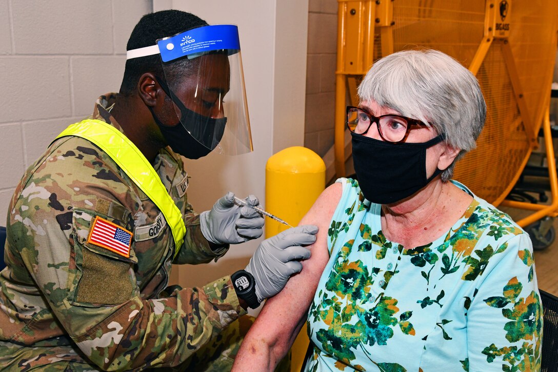 A soldier wearing a face mask and gloves injects a vaccine to an elderly patient wearing a face mask.