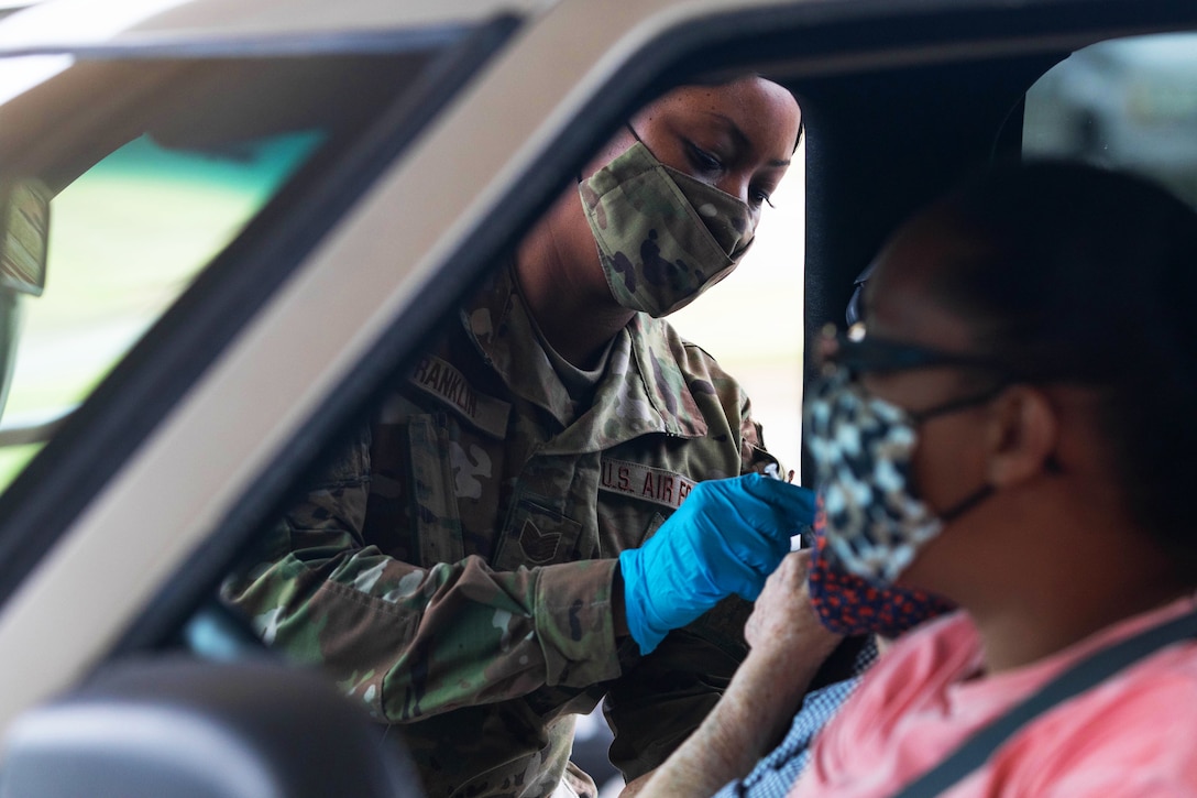 A soldier wearing a face mask and gloves reaches through the window of a vehicle to administer an injection to a passenger.