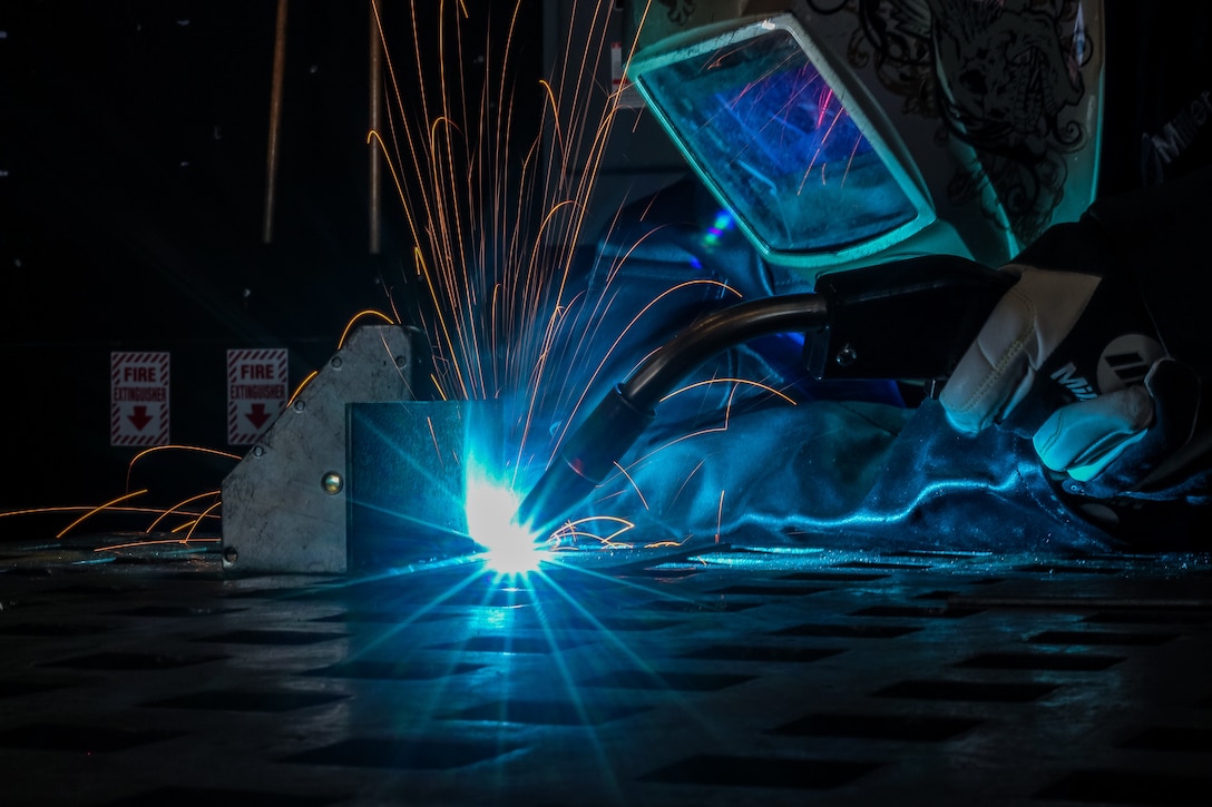 A Coast Guardsman welds as sparks fly illuminated by a blue light.