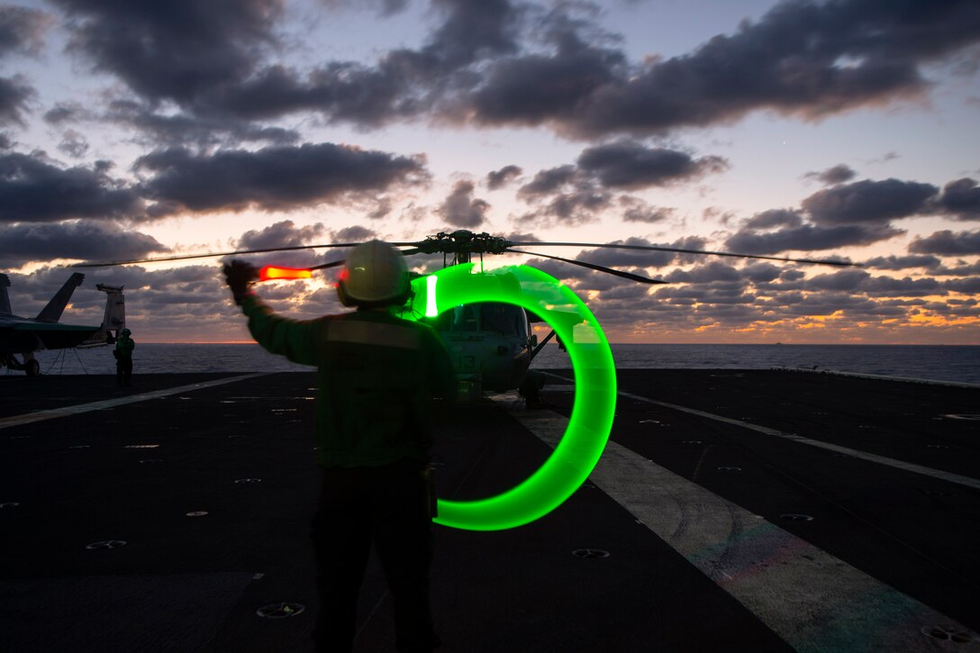 A sailor directs a helicopter on a ship while holding neon safety wands.