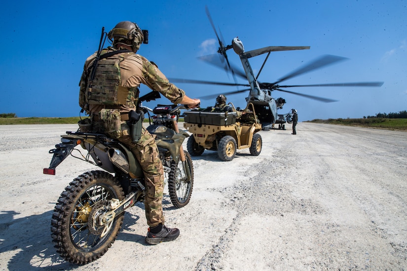 A service member sits on a motorcycle facing a helicopter.