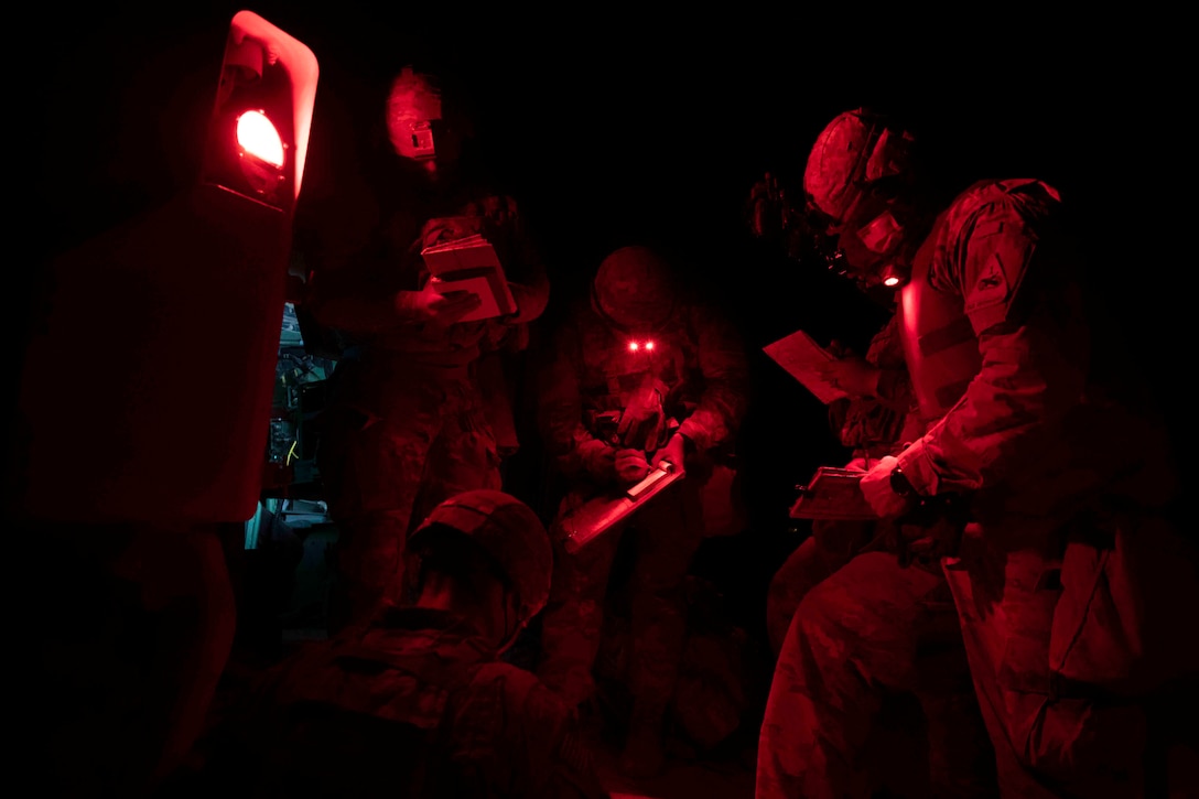 Soldiers illuminated by red light stand in a circle looking at papers.