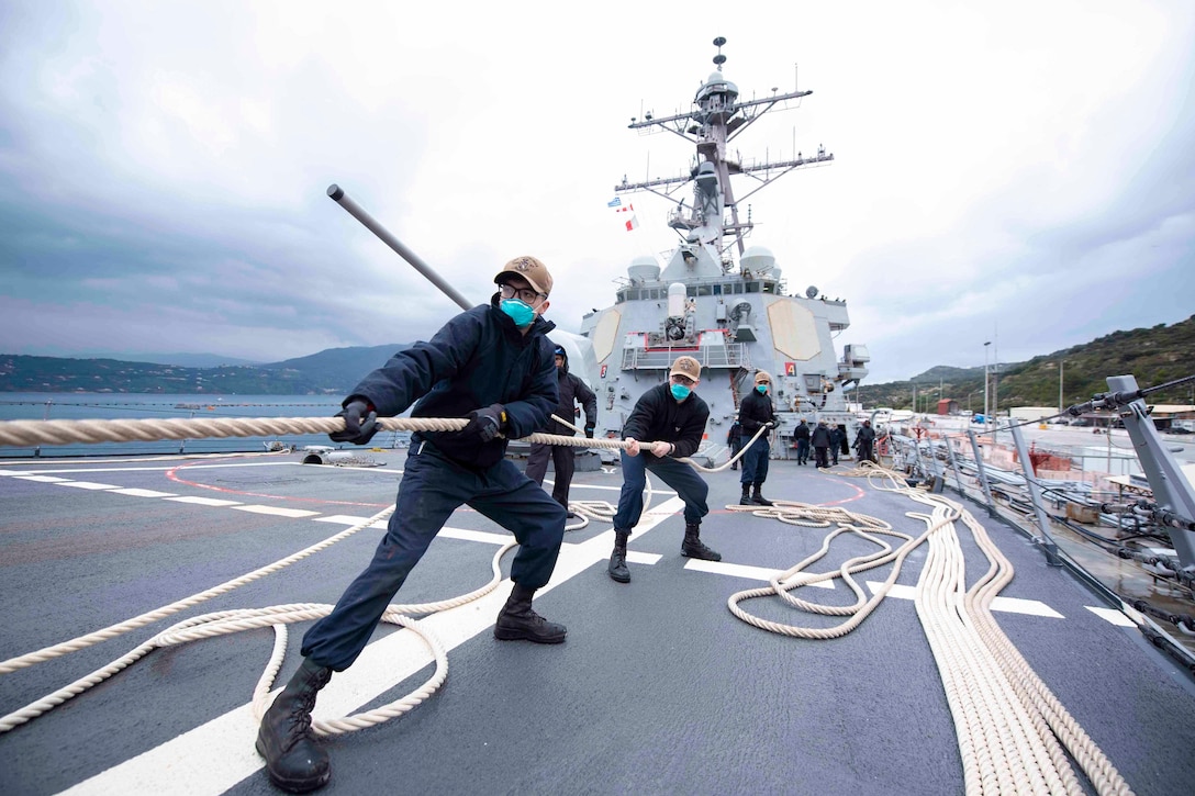 Sailors in a line pull a rope on the deck of a ship.