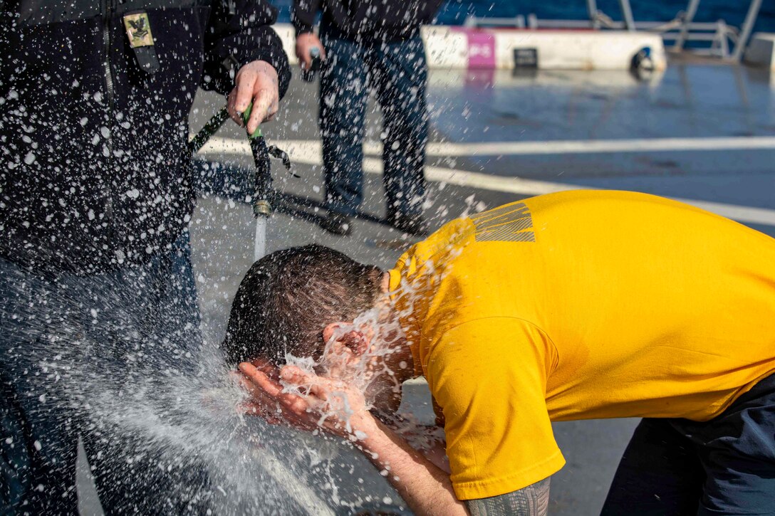 A service member sprays water on a sailor’s face on a ship.