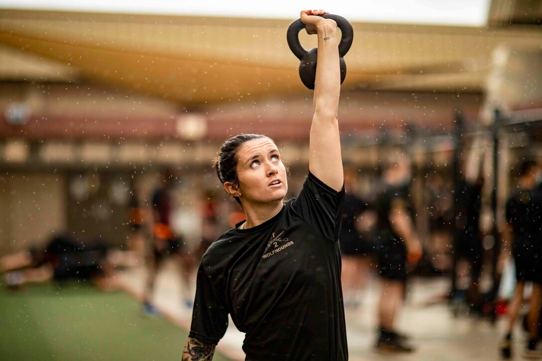 A soldier holds a kettlebell over her head as rain falls.