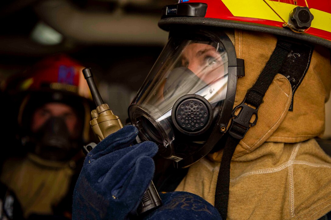 A sailor wearing fire protection gear talks into a radio.