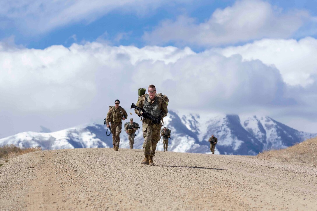Soldiers march on a dirt road while holding weapons; a mountain can be seen in the background.