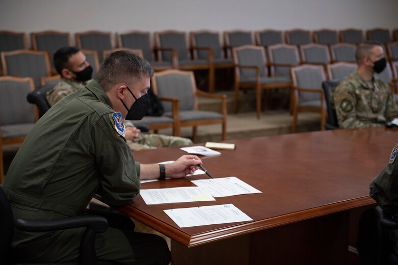A photo of three men sitting at a table with one of them looking at papers in front of him.