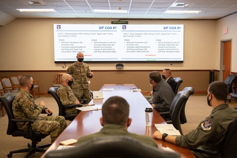 A photo of seven Airmen sitting at a table in front of a powerpoint slide.