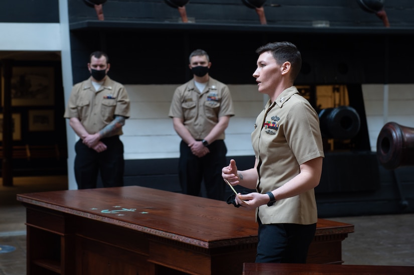 A sailor stands and speaks by a wood desk in a room.