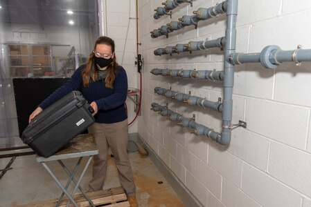 Photo of A woman stands in an industrial work area. The area appears to be a concrete chamber, with metal racks on the wall with spigots attached. The woman, wearing a mask and goggles for safety, is loading a black square case onto a base apparatus.