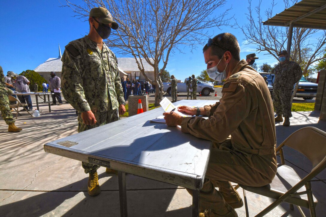 A sailor speaks to a fellow sailor sitting at a table while other service members stand in the background.